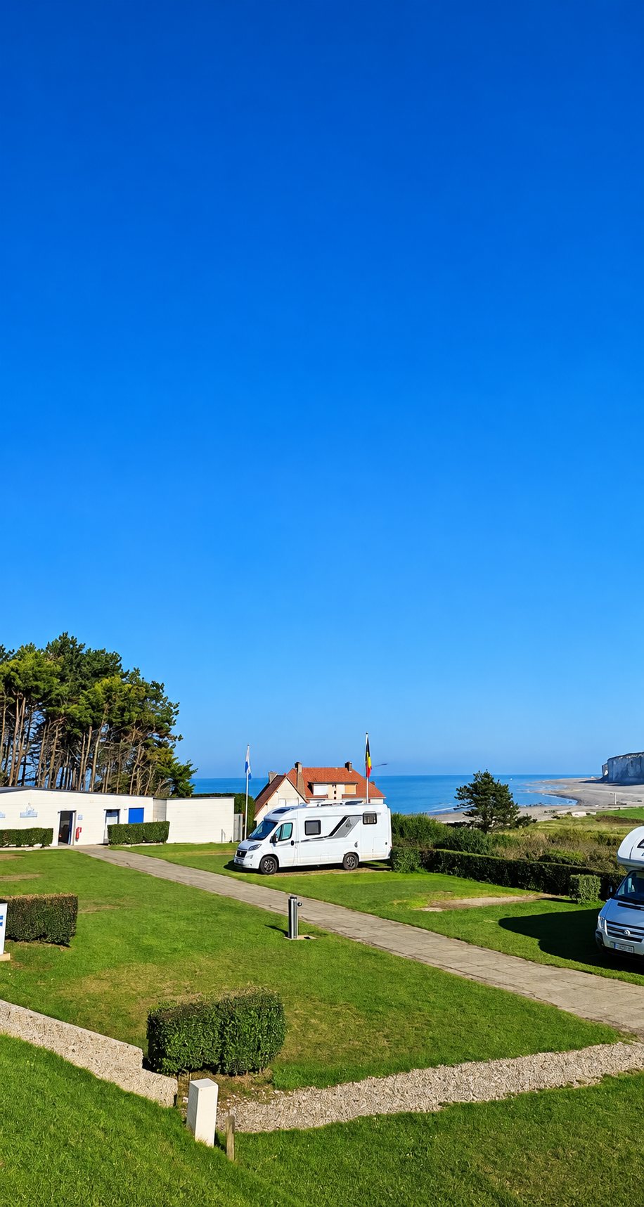 Vue depuis les hauteurs d'un camping herbeux avec des camping-cars garés, une maison au toit rouge et la mer au loin sous un ciel bleu.