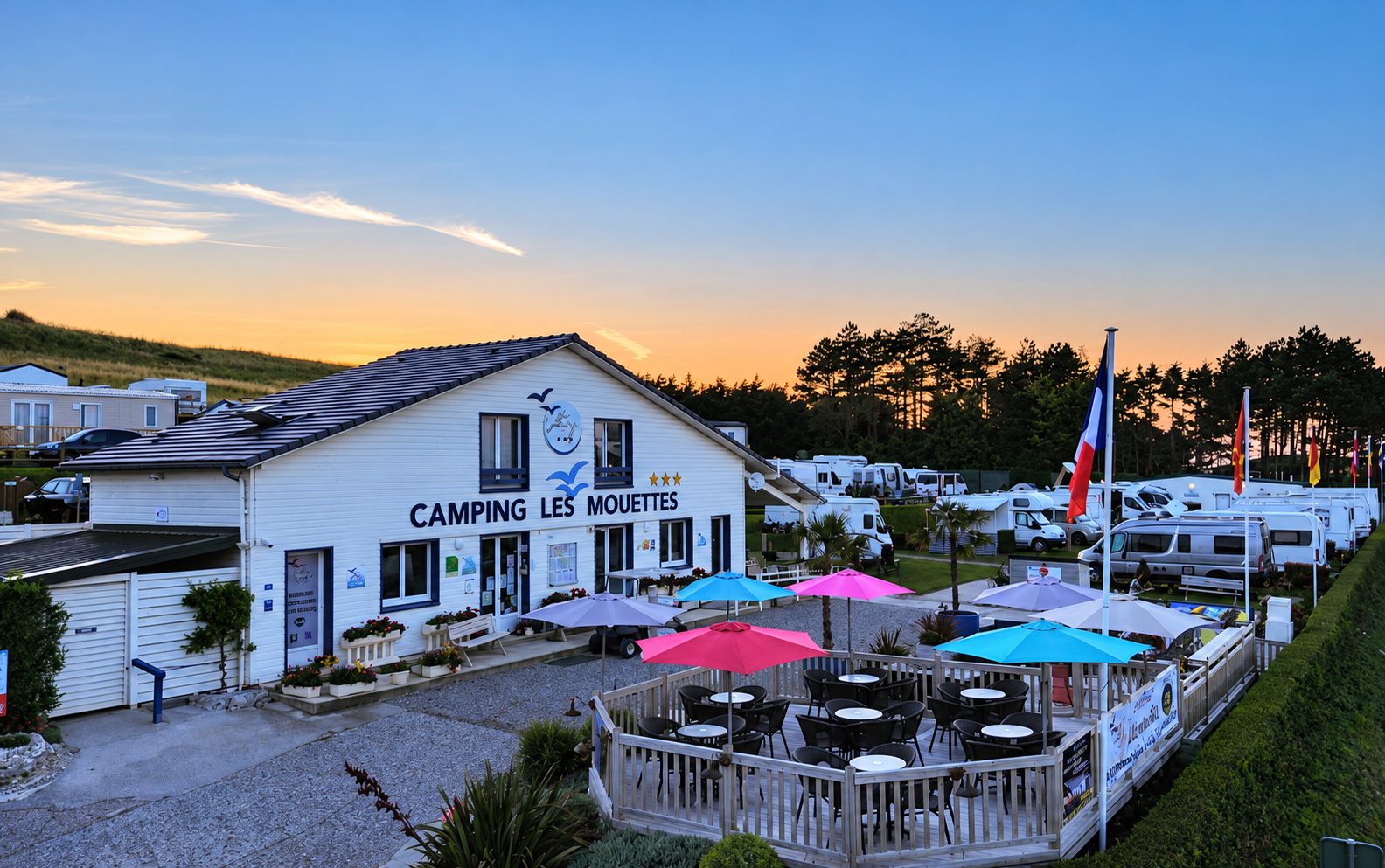 Au crépuscule, le bâtiment blanc du camping Les Mouettes donne sur une terrasse en bois avec parasols colorés et, derrière, des camping-cars garés sous les pins.