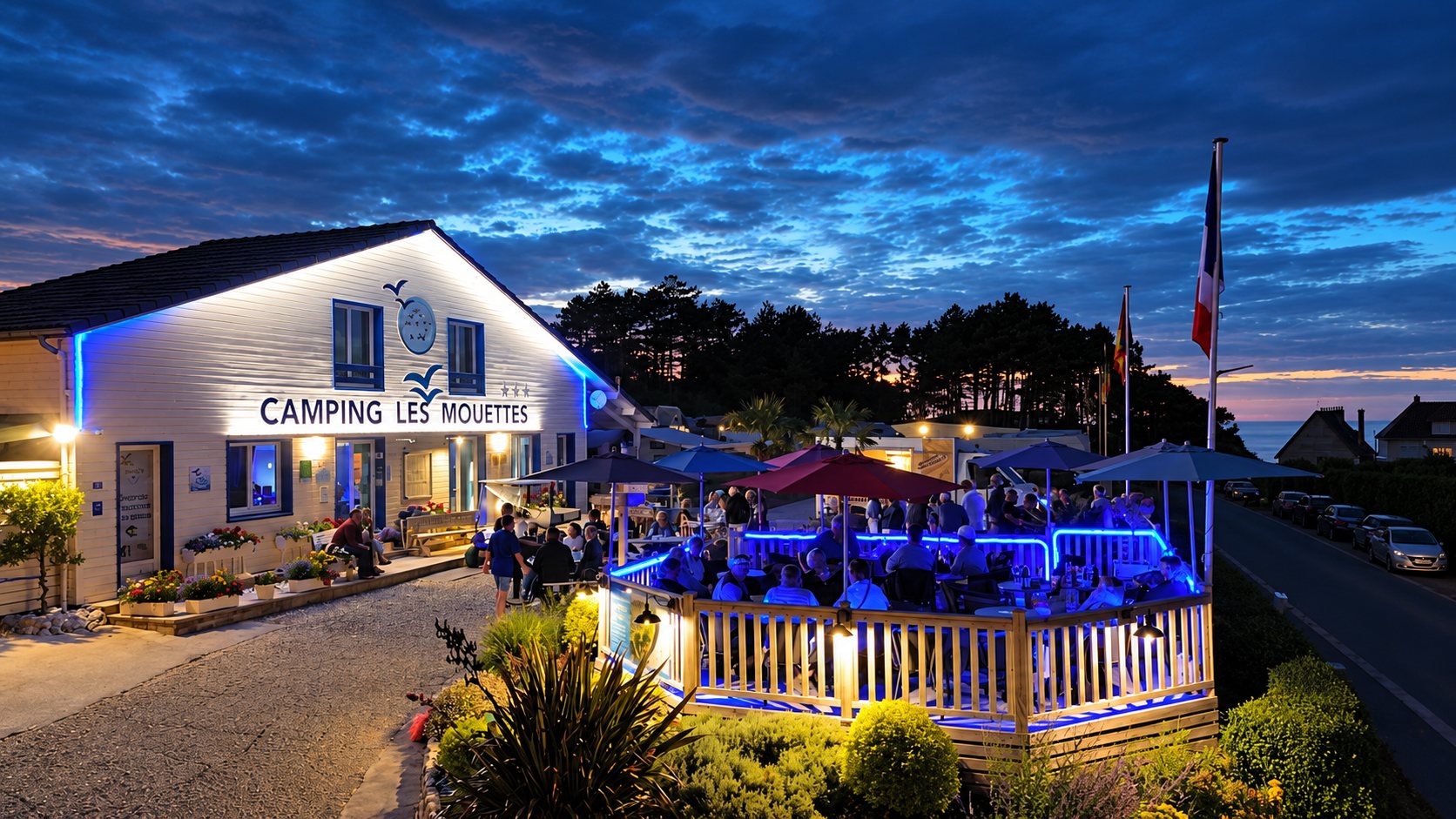 Au crépuscule, la façade du Camping Les Mouettes et sa terrasse remplie de clients sont illuminées de lumières bleues sous un ciel nuageux.