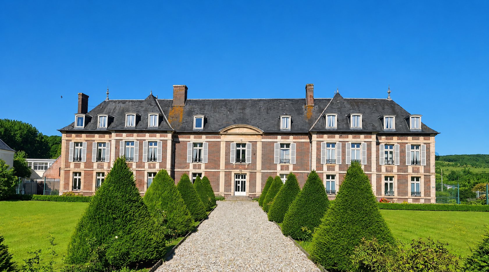 Façade d'un château en brique rouge et pierre, au toit d'ardoise, précédée d'une allée de gravier bordée d'ifs taillés en cône sous un ciel bleu.