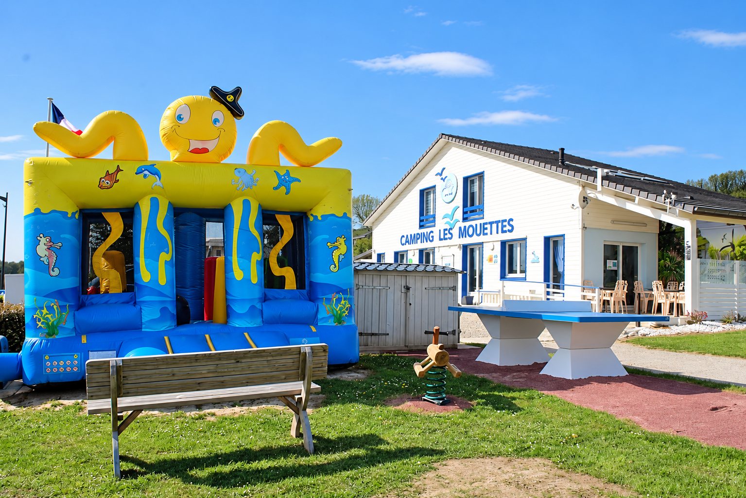 Un château gonflable en forme de pieuvre jaune et bleue, un banc et une table de ping-pong devant le bâtiment blanc du camping Les Mouettes.