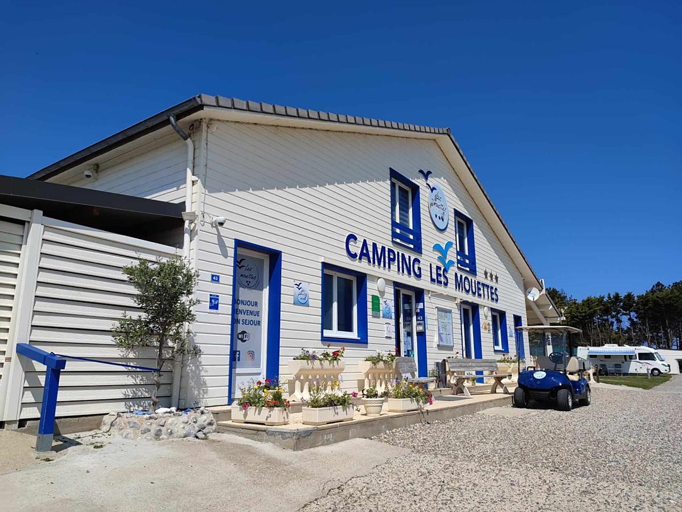 Bâtiment d'accueil blanc aux huisseries bleues du camping Les Mouettes, avec jardinières fleuries, voiturette de golf bleue et camping-car au fond sous un ciel dégagé.