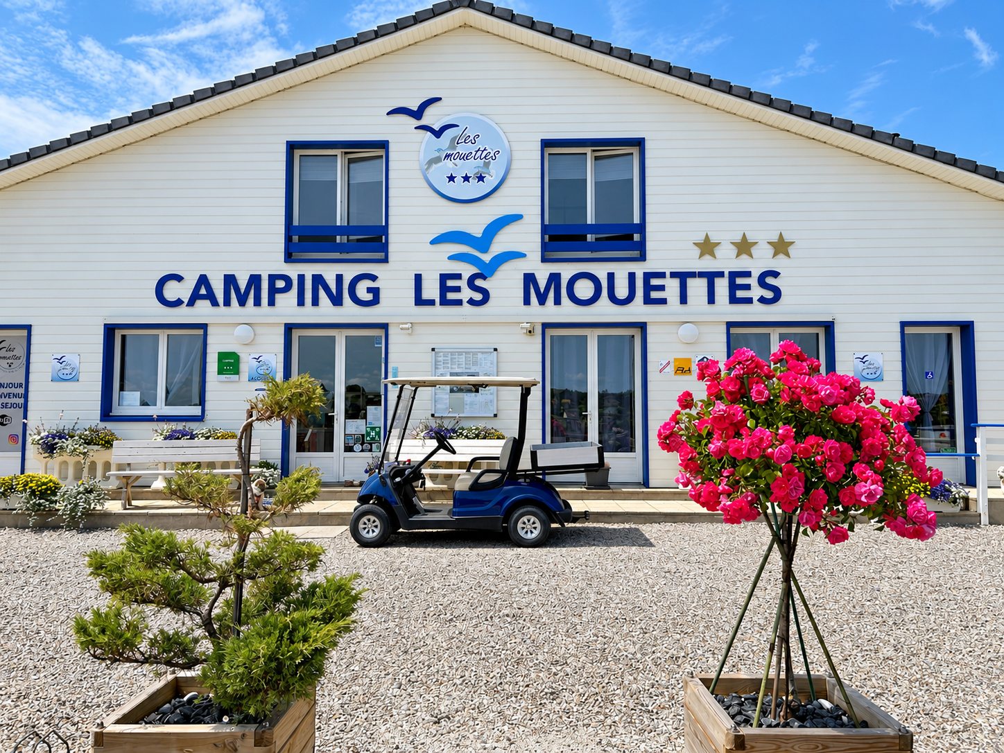 La façade blanche aux fenêtres bleues du camping Les Mouettes, avec une voiturette de golf bleue garée devant et des rosiers roses en pot au premier plan.