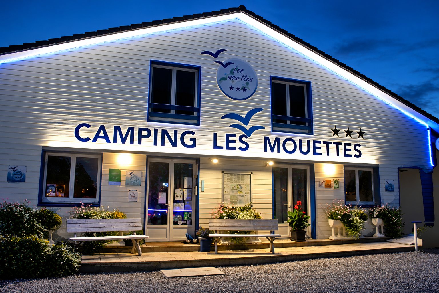 Façade en bois du camping Les Mouettes, éclairée à la tombée de la nuit, avec son enseigne, ses trois étoiles, des bancs et des jardinières fleuries devant l'entrée.