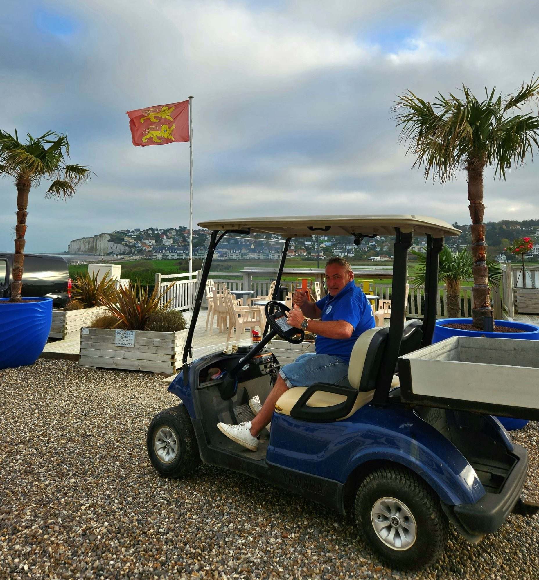Un homme en polo bleu fait un pouce levé au volant d'une voiturette électrique bleue, devant une terrasse en bois, des palmiers, un drapeau normand et les falaises au loin.