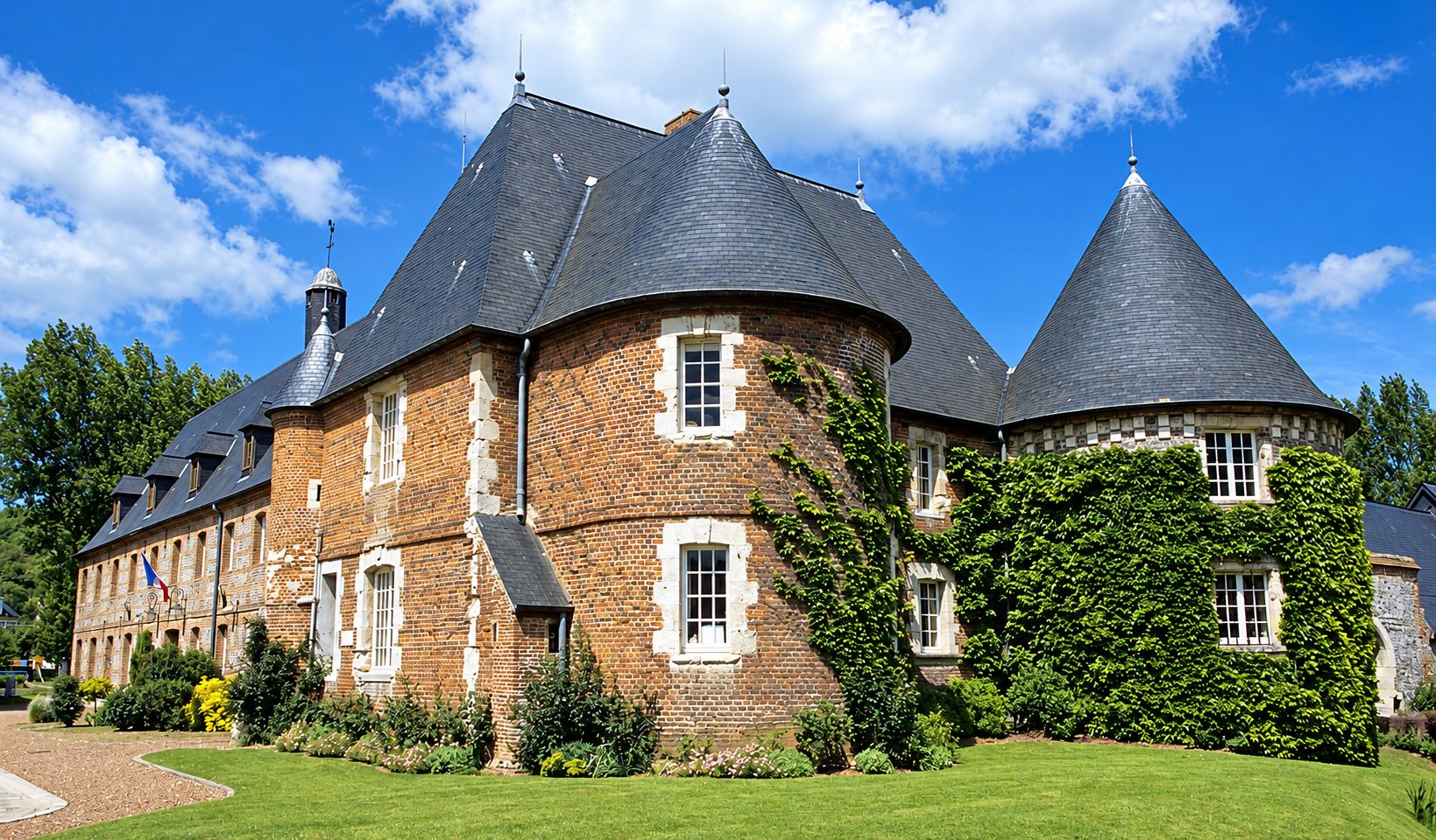 Un manoir en briques rouges et pierres blanches, avec deux tourelles aux toits d'ardoise, des murs en partie couverts de lierre et une pelouse au premier plan sous un ciel bleu.