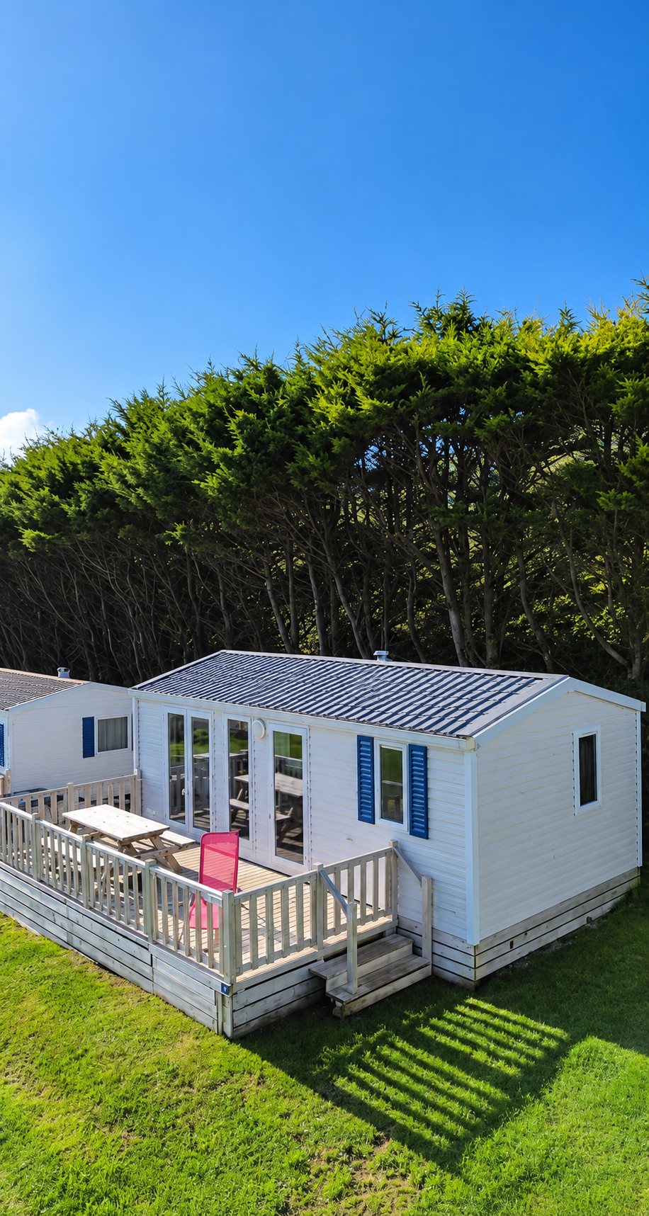 Un mobil-home blanc à volets bleus avec une terrasse en bois équipée d'une table et d'une chaise rouge, posé sur une pelouse devant une haie d'arbres.