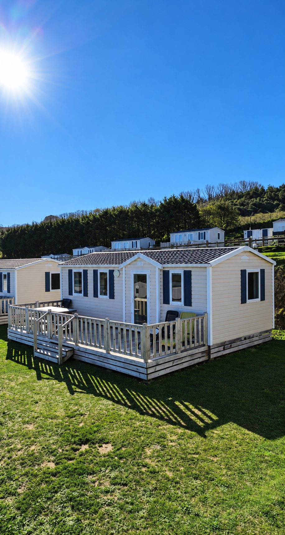 Un mobil-home beige aux volets bleu marine et à la terrasse en bois blanche, installé sur une pelouse verte sous un ciel bleu éclatant.