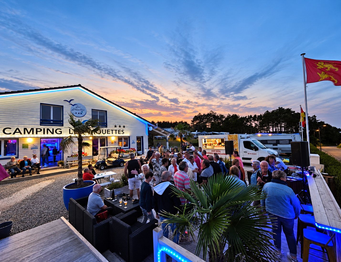 Au crépuscule, une foule de campeurs se rassemble sur une terrasse devant le bâtiment d'accueil illuminé d'un camping, près d'un camion de hot dogs.