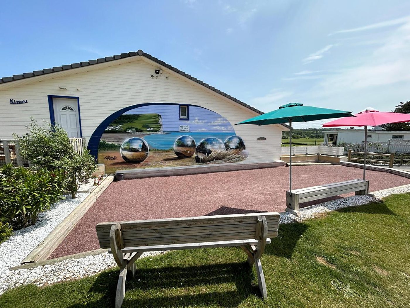 Un terrain de pétanque en gravier rouge avec des bancs en bois et des parasols, devant un chalet blanc orné d'une fresque de boules chromées sur fond de bord de mer.