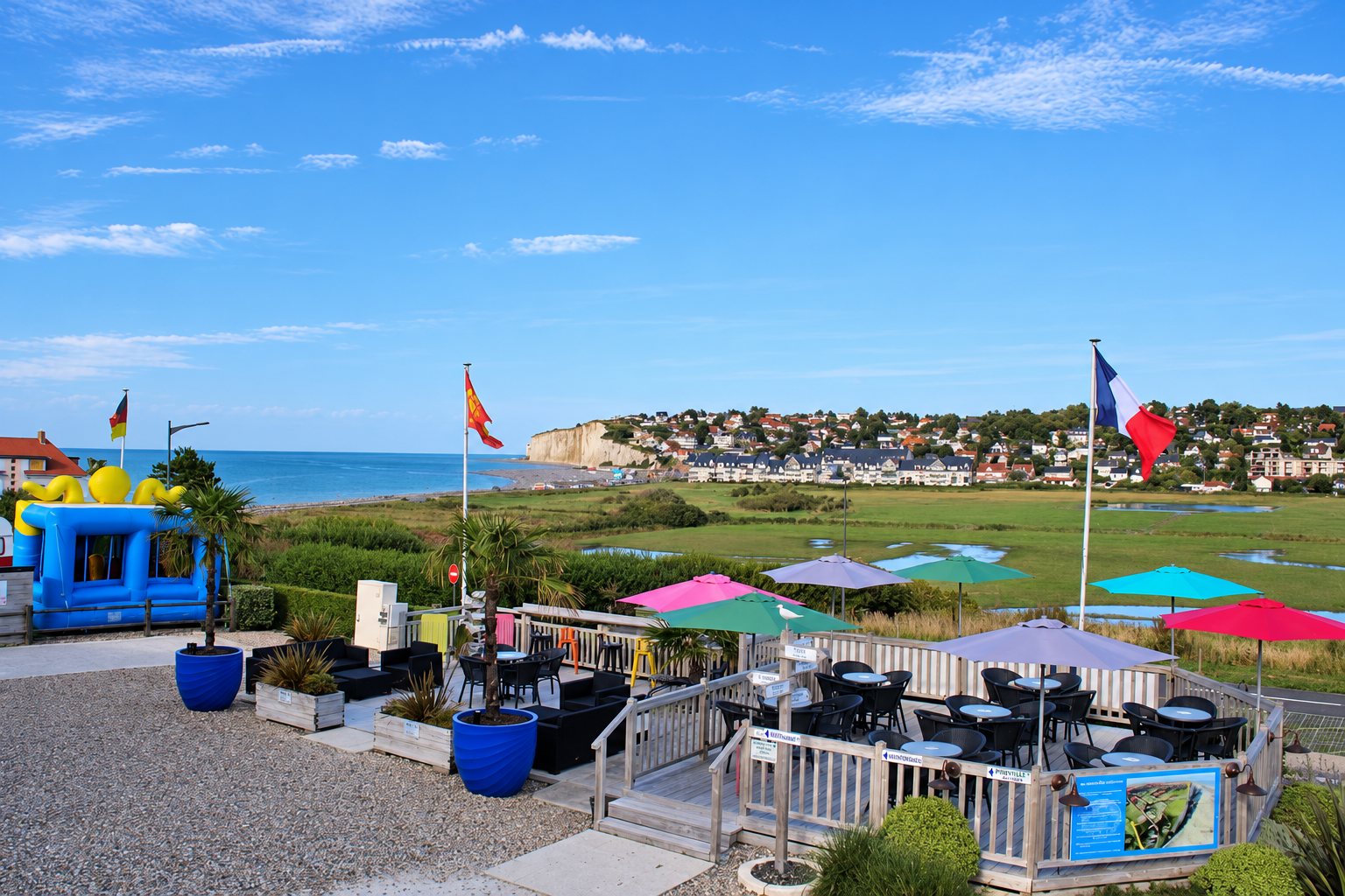 Une terrasse en bois garnie de tables, de chaises et de parasols colorés domine la mer, les prairies et une falaise blanche, avec un drapeau français et un château gonflable au premier plan.