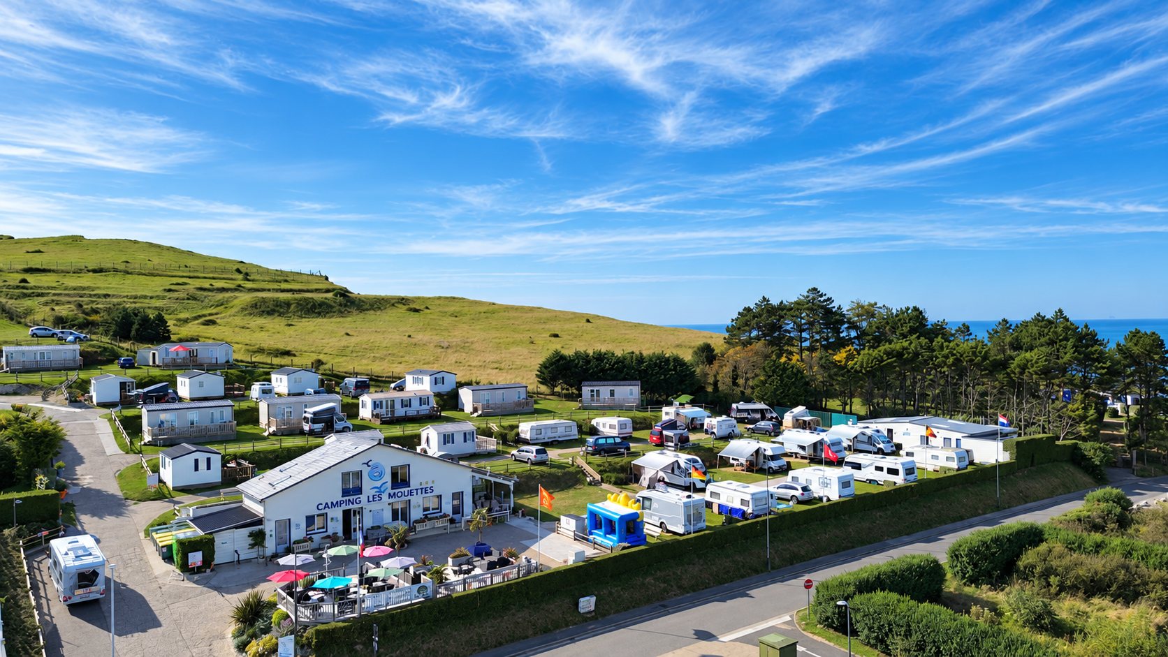 Vue aérienne d'un camping en bord de mer avec mobil-homes, caravanes et camping-cars, au pied d'une colline verte sous un grand ciel bleu.